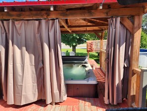 Un bain à remous sous une pergola en bois, agrémentée de rideaux, entouré d’une terrasse en bois et d’un arbre.
