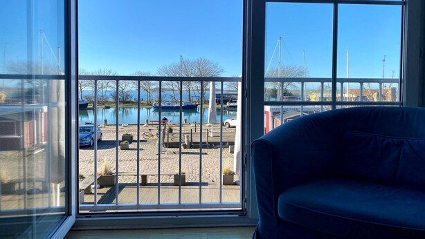 A room with a large window, a blue sofa, and a view of a dock with boats and a body of water. - Ferienwohnung in Orth auf Fehmarn (Orth)
