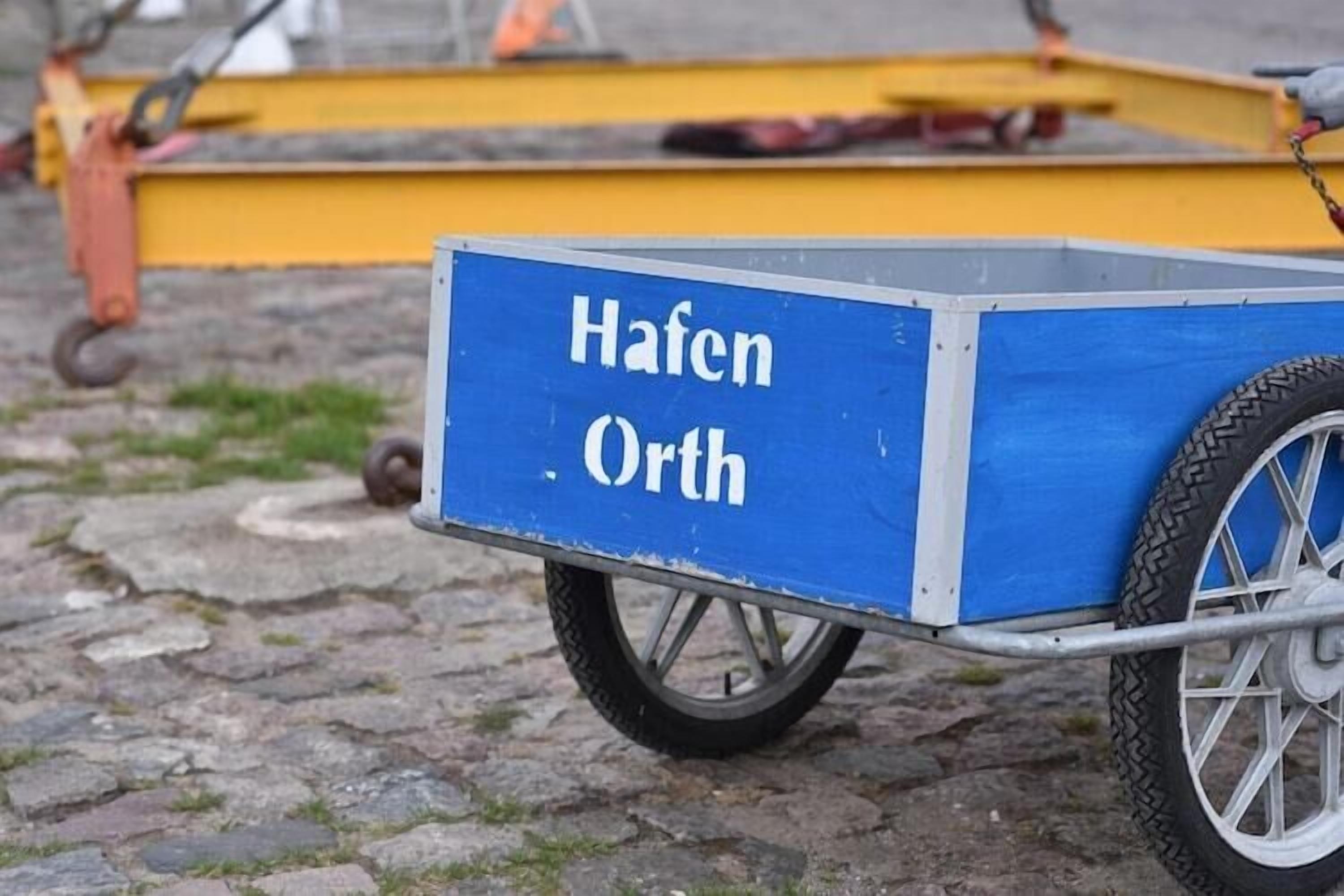 A blue cart with 'Hafen Orth' written on it, positioned on cobblestone ground.