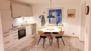 Ett modernt kök med vita skåp, en inbyggd ugn, ett matbord med stolar, en hänglampa och ett fönster med utsikt.