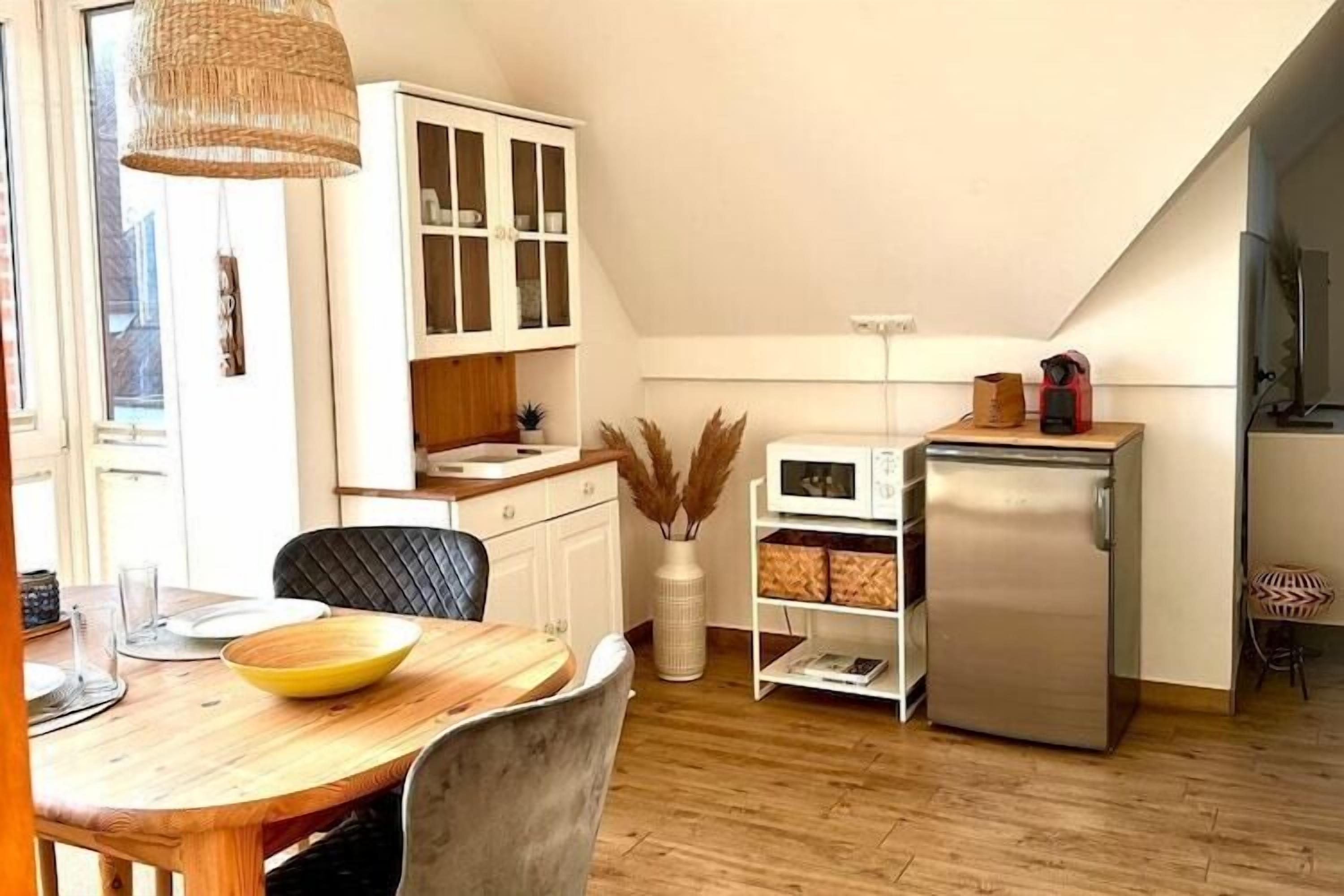 Una cocina con mesa de comedor, sillas, microondas, refrigerador, armarios y una lámpara colgante.