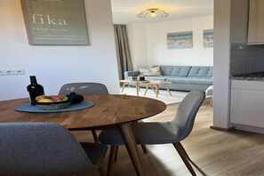A modern living room with a sofa, a dining table with chairs, a kitchen area, and a wall-mounted artwork. - Ferienwohnung in Lemkenhafen auf Fehmarn (Lemkenhafen)