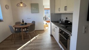 Uma cozinha compacta com área de jantar, com uma mesa de madeira, cadeiras, forno embutido e luminária suspensa.