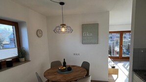 Uma sala de jantar com uma mesa redonda de madeira, quatro cadeiras, um relógio de parede, uma luminária pendente geométrica e uma moldura com uma frase na parede.