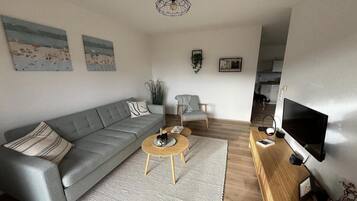 Een woonkamer met een grijze bank, een klein rond salontafeltje, een televisie en een boekenplank.