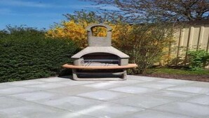 Ein Stein-Pizzaofen im Freien mit Schornstein, auf einer gepflasterten Terrasse, umgeben von viel Grün.