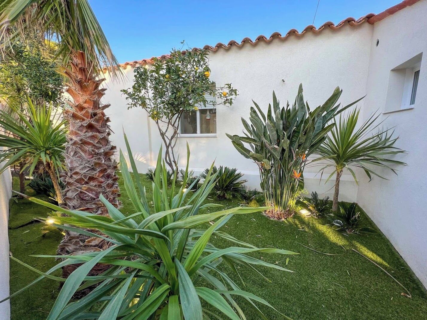 A white building with a tiled roof, surrounded by various plants and a small window.