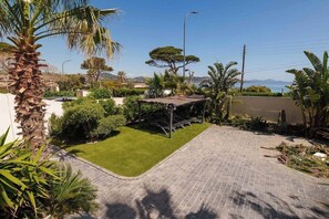 Een verhard terras met een klein gazon, een pergola en diverse tropische planten.