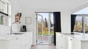 A modern kitchen with white cabinetry, a stainless steel sink, and a coffee maker. There is a sliding glass door leading to an outdoor area. - 4 Star Holiday Home in Rude (Bisserup)