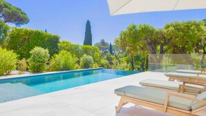 Une piscine entourée de verdure, avec des chaises longues et un parasol, sous un ciel dégagé.