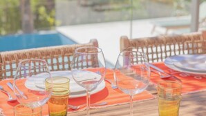 Une table à manger avec des verres à vin et un verre d’eau, placée sur une terrasse extérieure meublée de mobilier en osier, offrant une vue sur des arbres et une piscine.