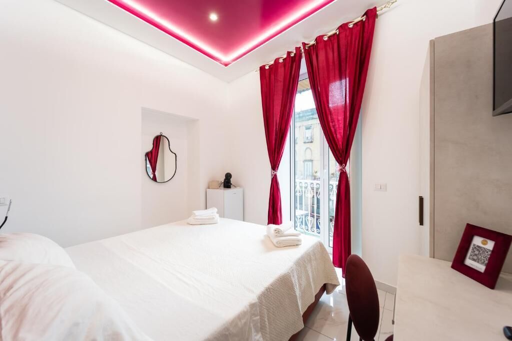 Une chambre d’hôtel avec un lit, une table de chevet, un miroir, une fenêtre avec des rideaux rouges et une vue sur les bâtiments environnants.