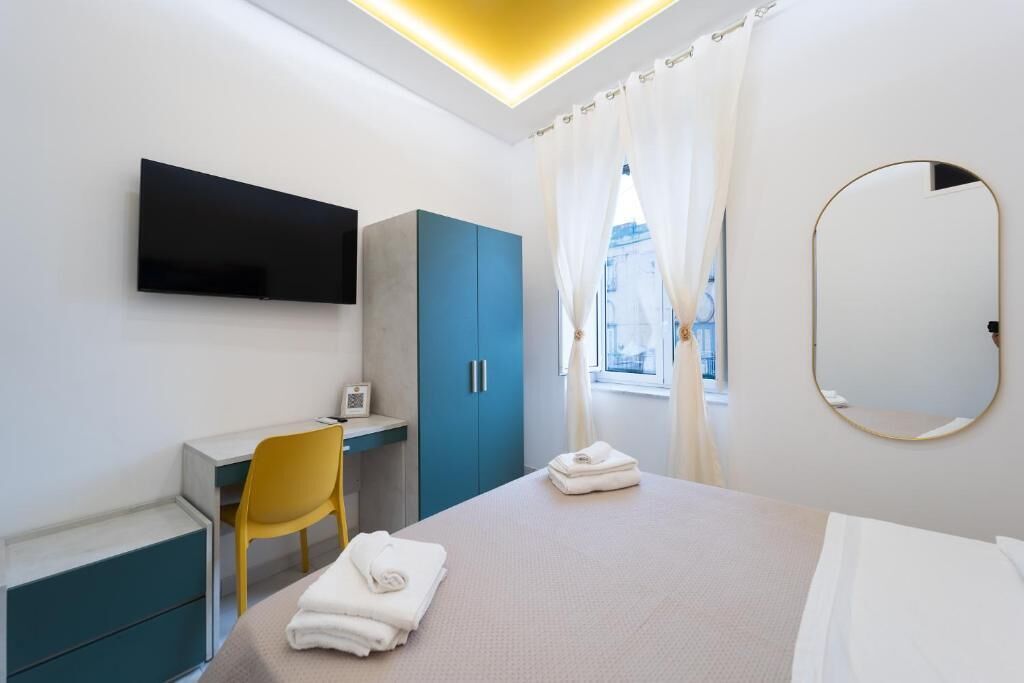 A modern hotel room with a bed, a desk, a chair, a TV, a wardrobe, and a window with curtains.