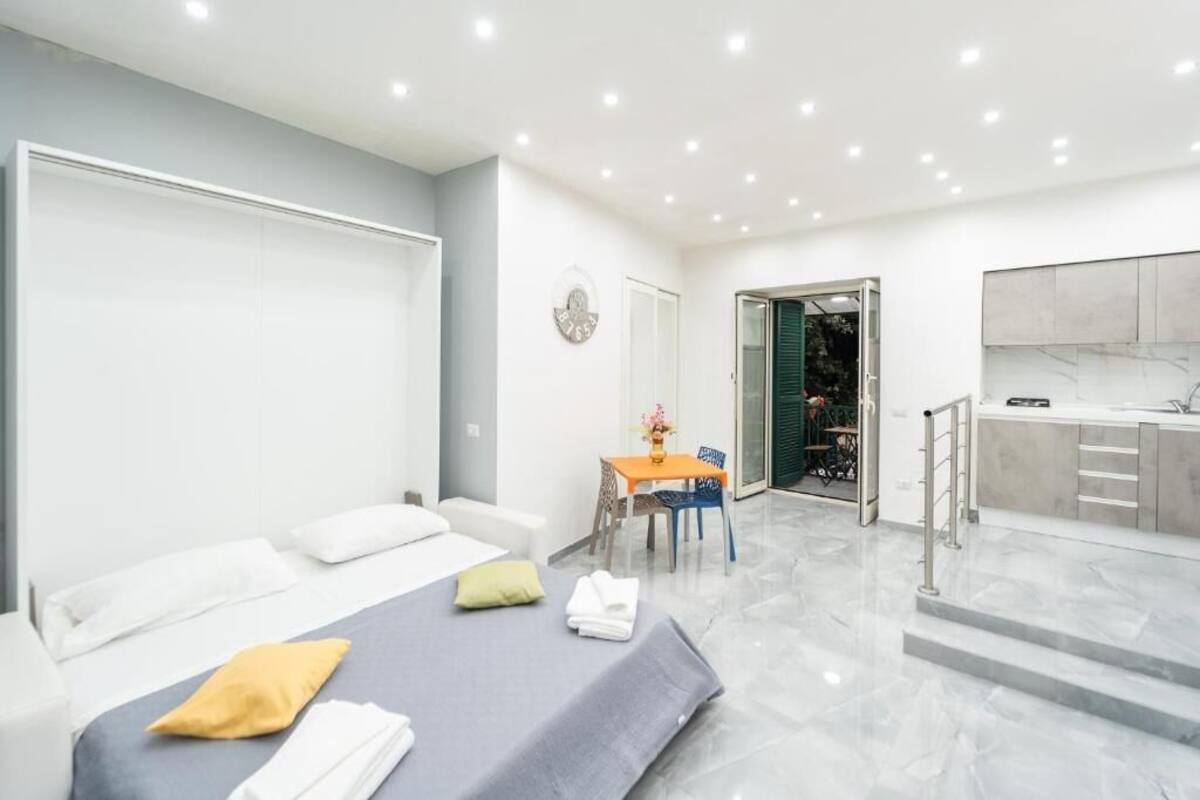 Quarto de hotel moderno com cama, uma mesinha com cadeiras, banheiro e cozinha.
