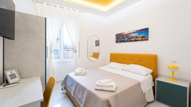 A modern hotel room with a bed, a desk, a chair, a bedside table, a lamp, a painting, and a window with curtains. - Vico Teresa 148 (Naples)