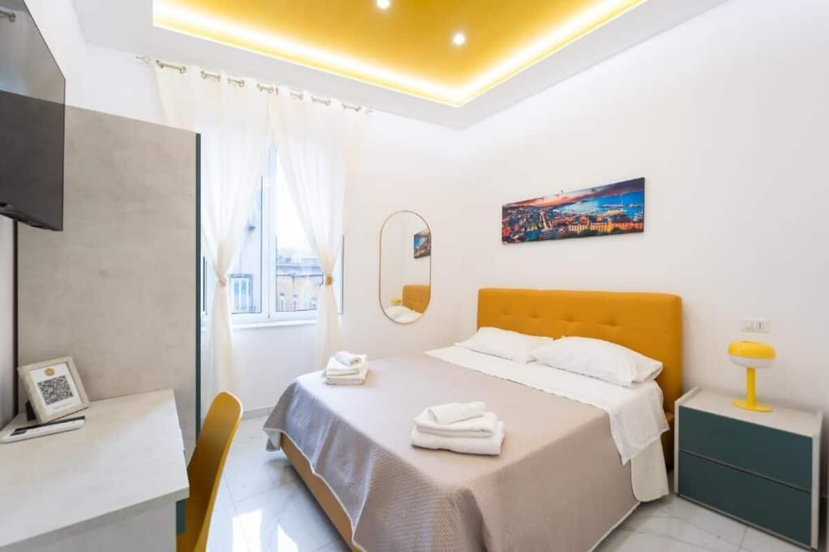Quarto de hotel moderno com cama, escrivaninha, cadeira, mesa de cabeceira, abajur, um quadro e janela com cortinas.