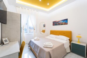 A modern hotel room with a bed, a desk, a chair, a bedside table, a lamp, a painting, and a window with curtains. - Vico Teresa 148 (Naples)