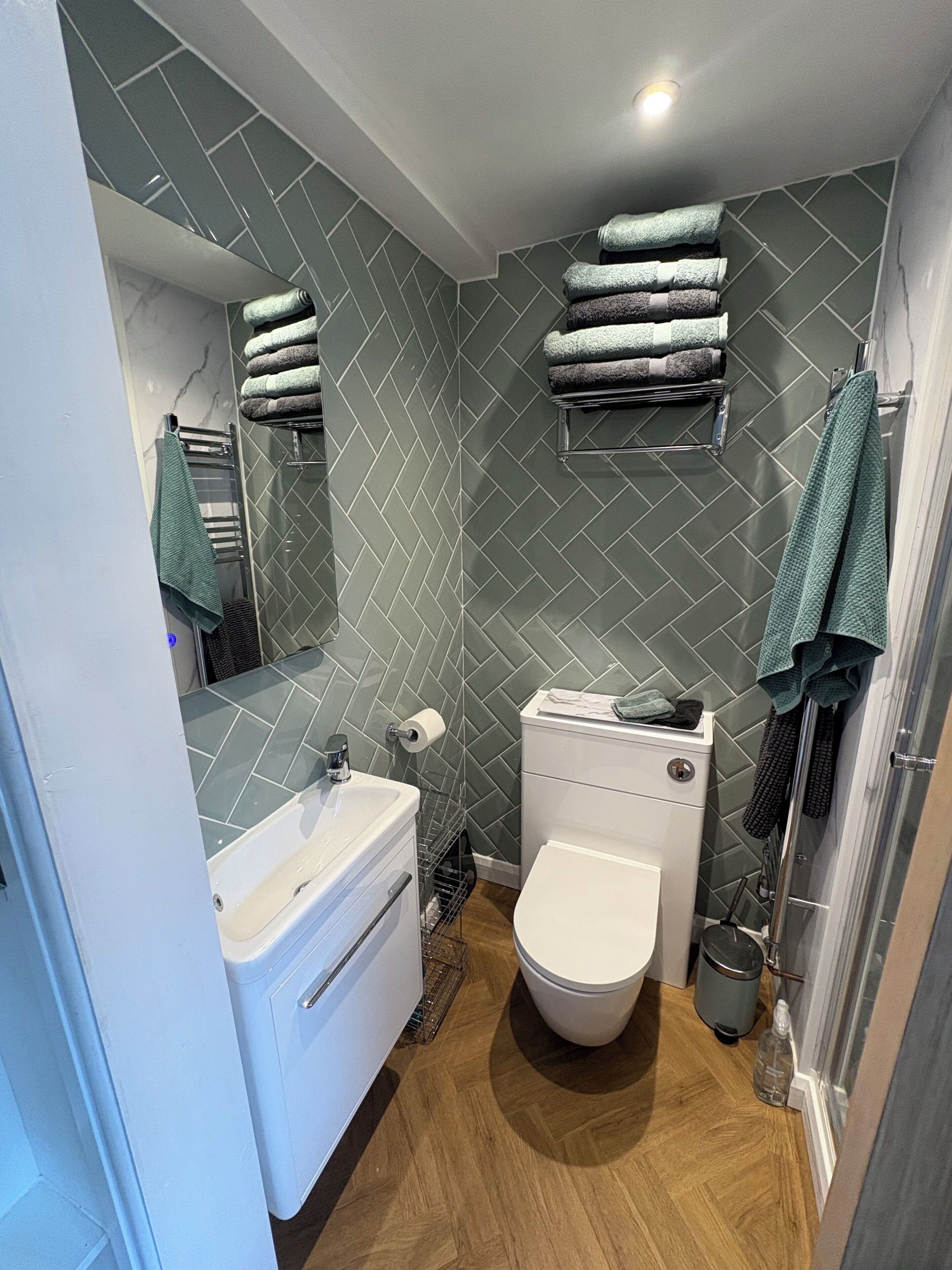 Une salle de bain avec un lavabo, des toilettes et une douche, le tout de couleur blanche. Des serviettes sont pliées et empilées sur une étagère, et un miroir est placé au-dessus du lavabo.