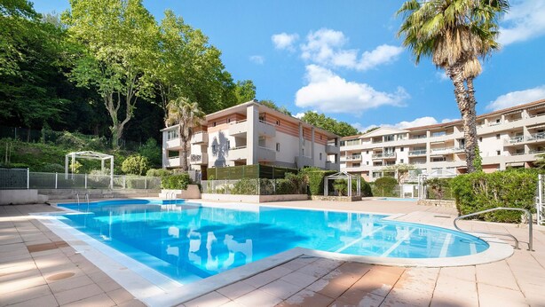 Une piscine entourée d’une terrasse carrelée, un immeuble à plusieurs étages doté de balcons, et une végétation luxuriante en arrière-plan.
