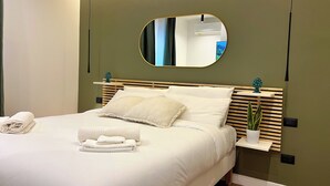 Un lit bien fait, avec du linge de lit blanc et des oreillers, une table de chevet agrémentée d’une plante et un miroir fixé au mur.