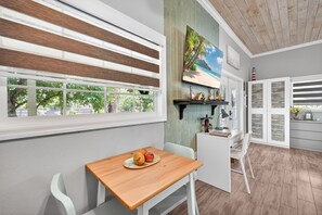 A dining area with a wooden table and chairs, a window with blinds, a painting of a beach scene, and a kitchen area with cabinets and a sink. - Adorable Estero Cottage - Heated Pool/Spa - Sauna (Estero)