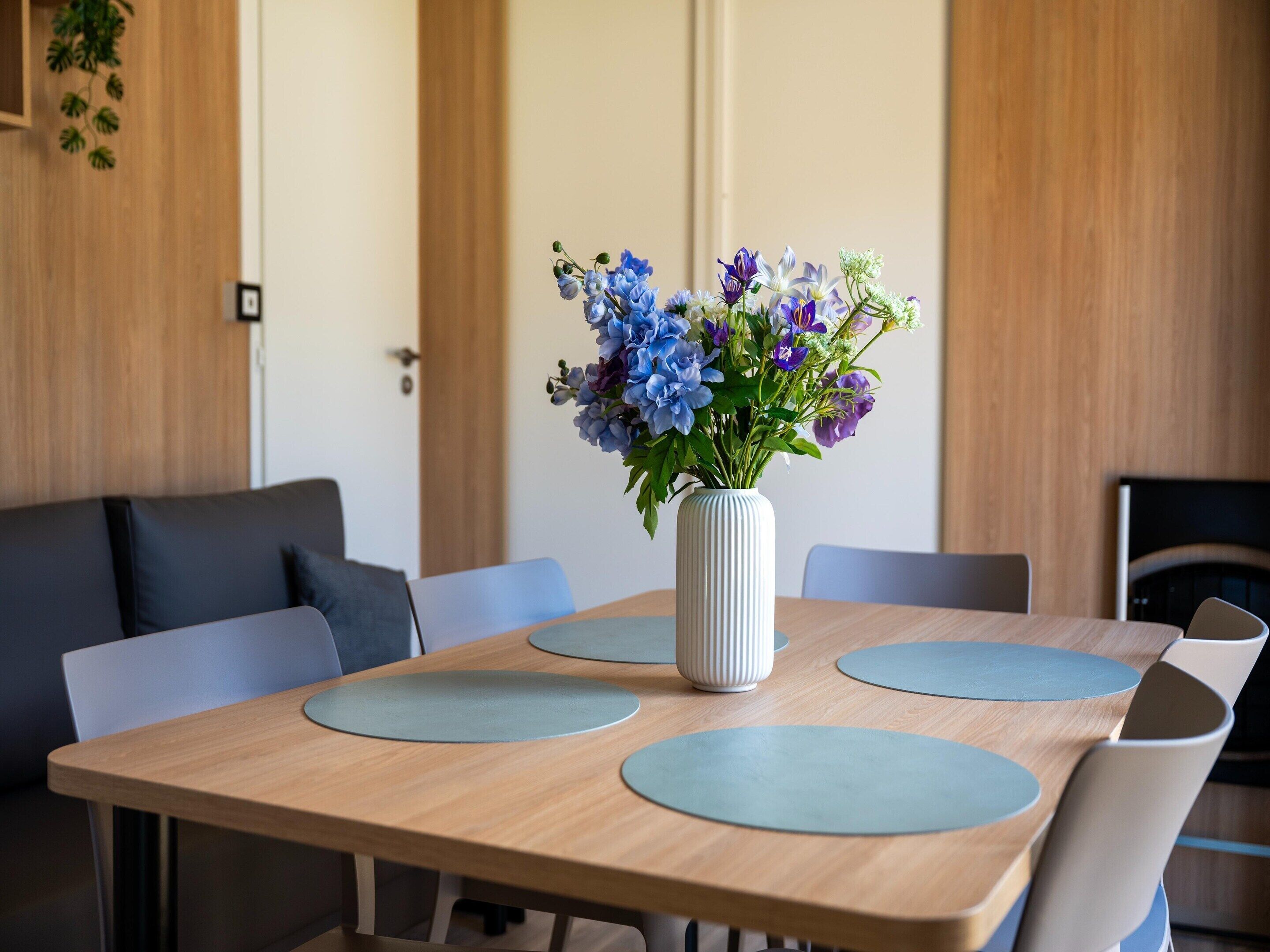 Una mesa de comedor de madera con un jarrón blanco lleno de flores, rodeada de sillas azules.