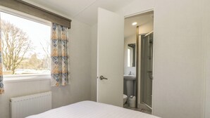 A bedroom with a bed, a window with curtains, a door leading to a bathroom, and a radiator. - Standard Caravan at Three Lochs Holiday Park (Newton Stewart)