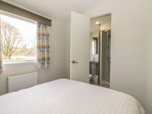 A bedroom with a bed, a window with curtains, a door leading to a bathroom, and a radiator. - Standard Caravan at Three Lochs Holiday Park (Newton Stewart)