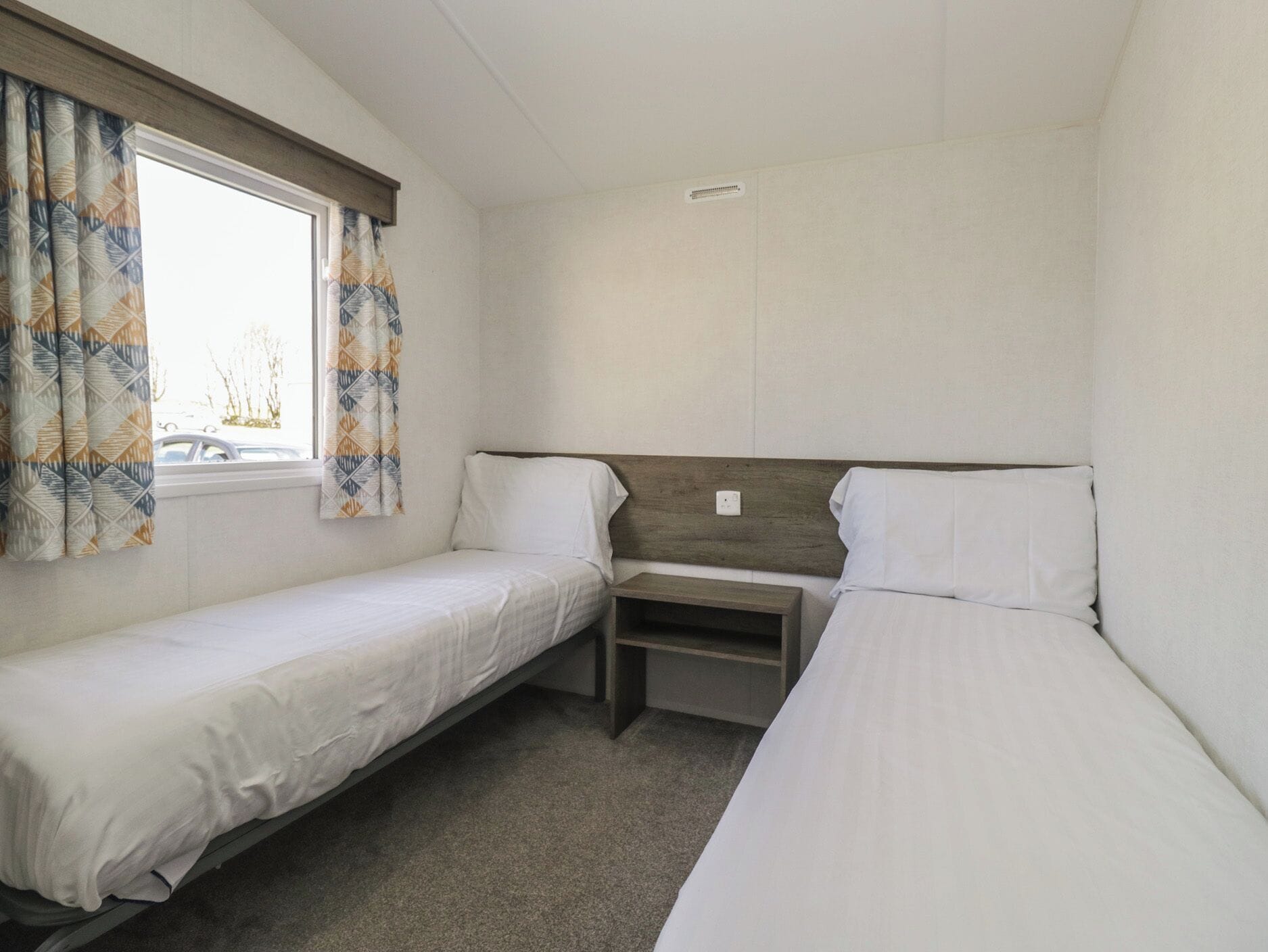 A small, single-room accommodation with two beds, a window with curtains, a wooden headboard, and a built-in shelf.