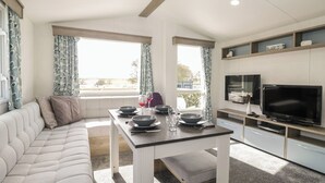 A modern living room with a sofa, a dining table with bowls and glasses, a flat-screen TV, and a kitchen area with cabinets and a countertop. - Standard Caravan at Three Lochs Holiday Park (Newton Stewart)