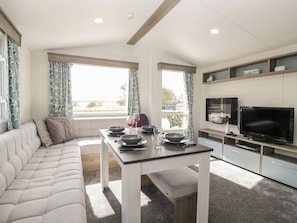 A modern living room with a sofa, a dining table with bowls and glasses, a flat-screen TV, and a kitchen area with cabinets and a countertop. - Standard Caravan at Three Lochs Holiday Park (Newton Stewart)