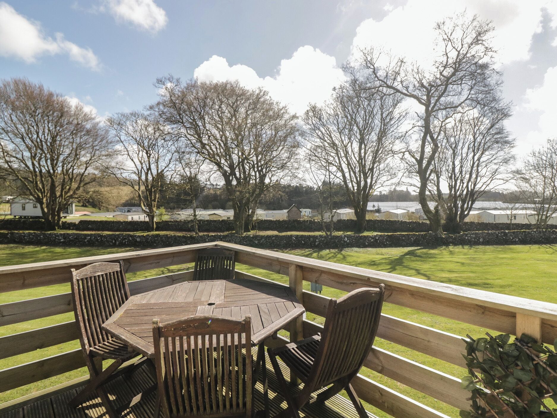 A wooden deck with a table and chairs, overlooking a grassy area with trees and distant buildings.