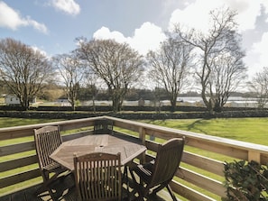 Une terrasse en bois avec une table et des chaises, donnant sur un espace verdoyant agrémenté d’arbres et bordé de bâtiments éloignés.
