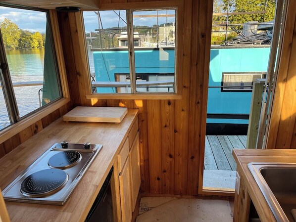 A compact kitchen with wooden walls, a window overlooking a dock, and a view of boats.