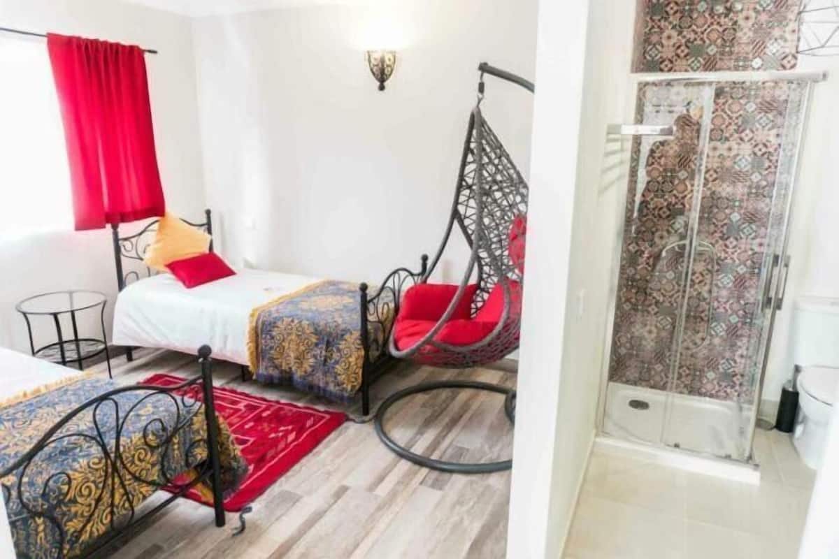 A room with a bed, a hanging chair, a small table, and a shower area.