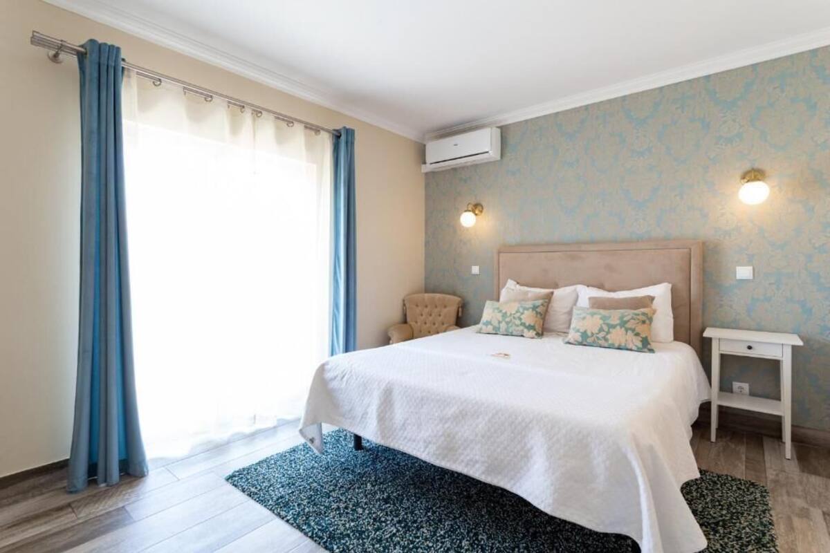 A bedroom with a bed, curtains, a nightstand, a chair, and an air conditioning unit.