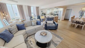 Un salon moderne avec un canapé, un fauteuil et une table basse. On aperçoit une cuisine et un coin repas à l’arrière-plan.