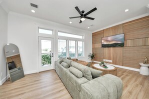 A modern living room with a large grey sofa, a ceiling fan, a flat-screen TV mounted on a wooden wall, and a large window with a view of the outdoors.