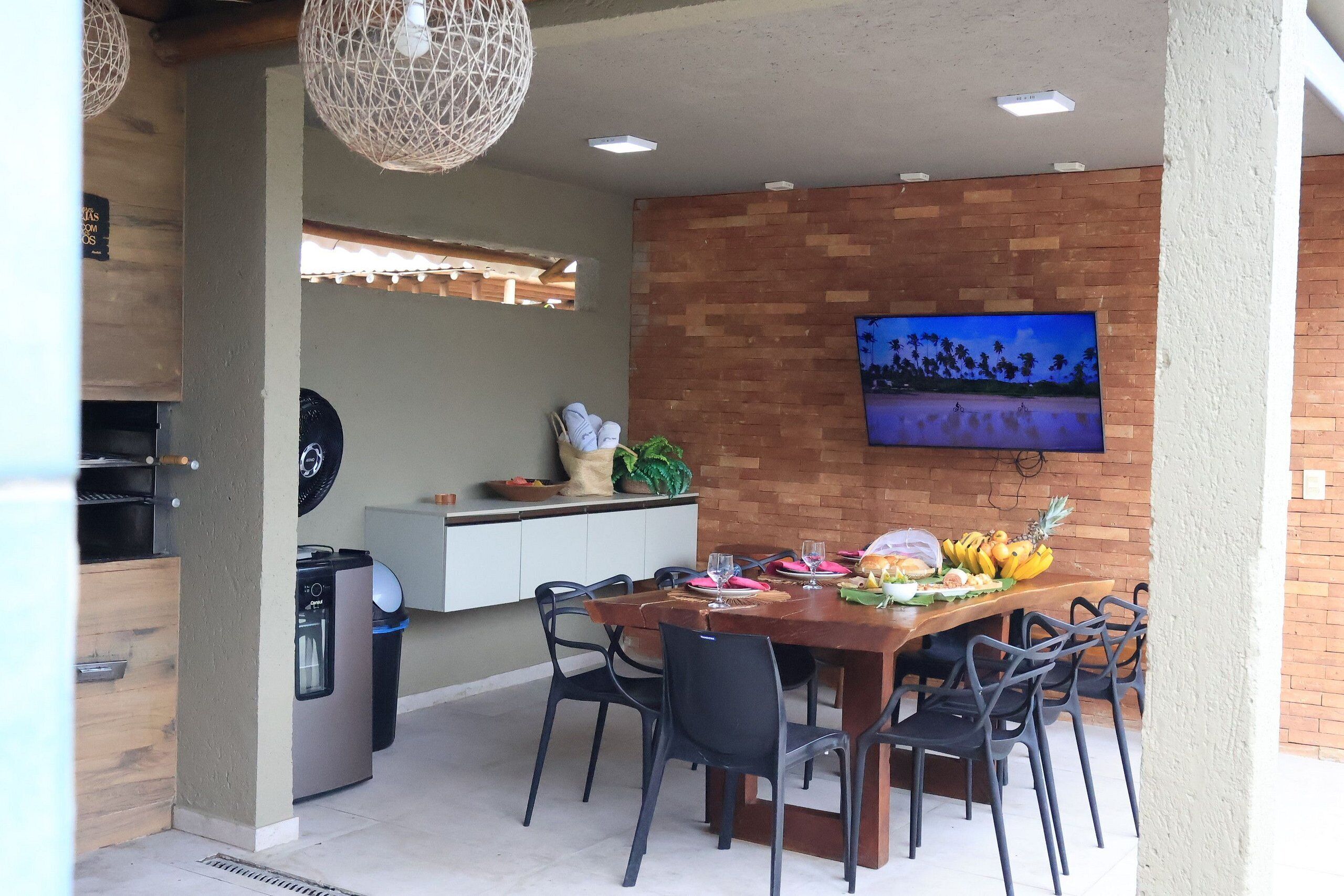 Un comedor con pared de ladrillo, un televisor montado sobre una mesa de madera, y una zona de cocina con una parrilla y dispensador de agua.