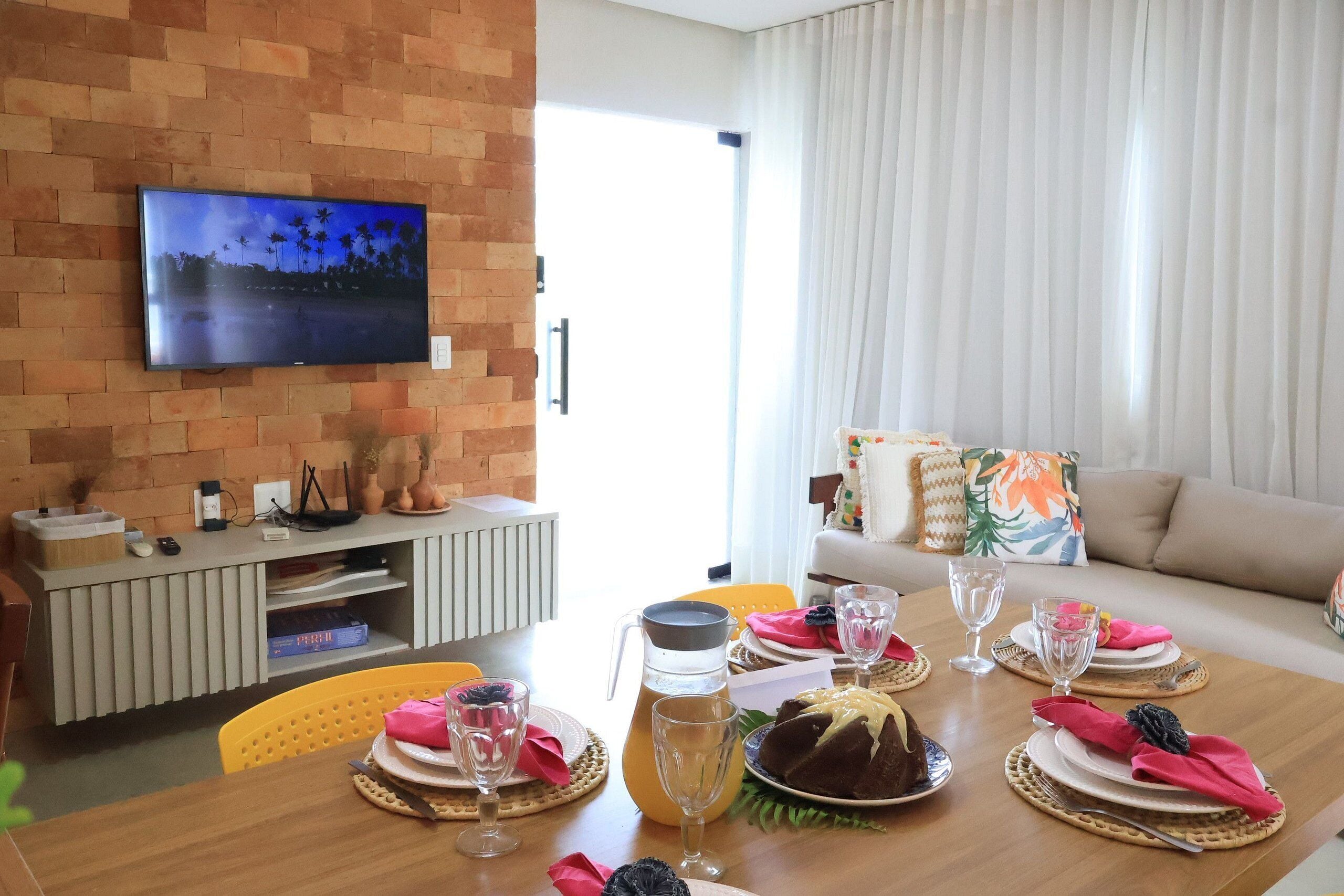Uma sala de jantar com uma mesa posta para quatro pessoas, uma televisão fixada em uma parede de tijolos e um sofá ao fundo.