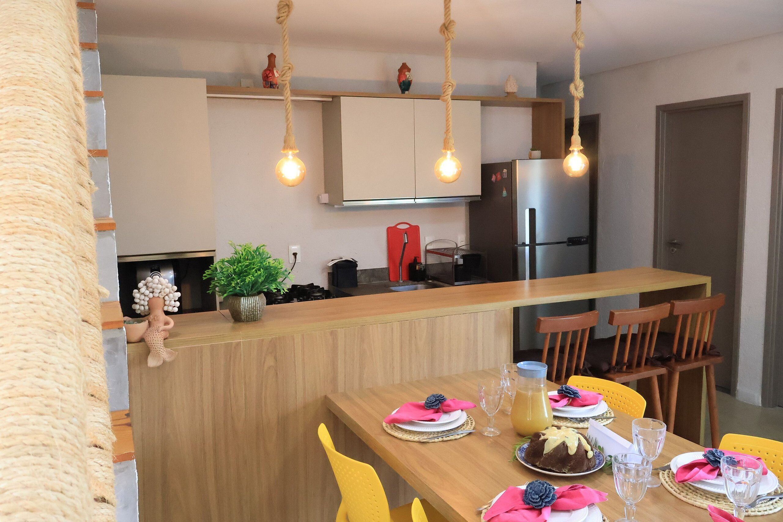 Una cocina moderna con armarios de madera, una isla integrada, lámparas colgantes y un comedor con sillas amarillas y una mesa de madera para dos.