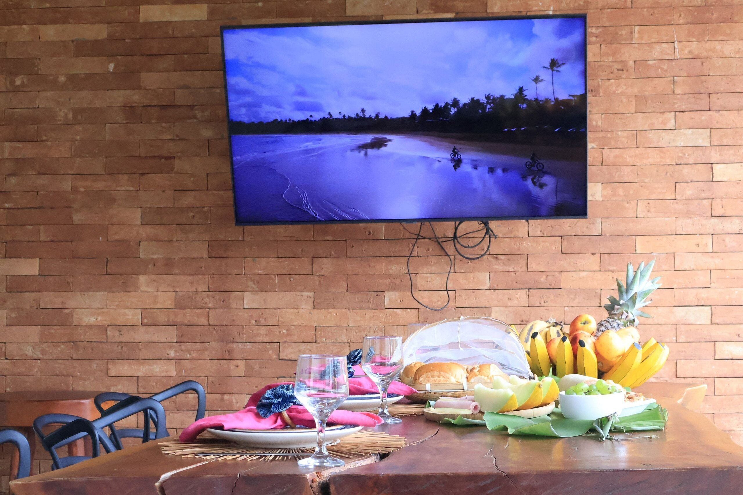 Una mesa de comedor con un televisor mostrando una escena de playa, un frutero y una cesta de pan.