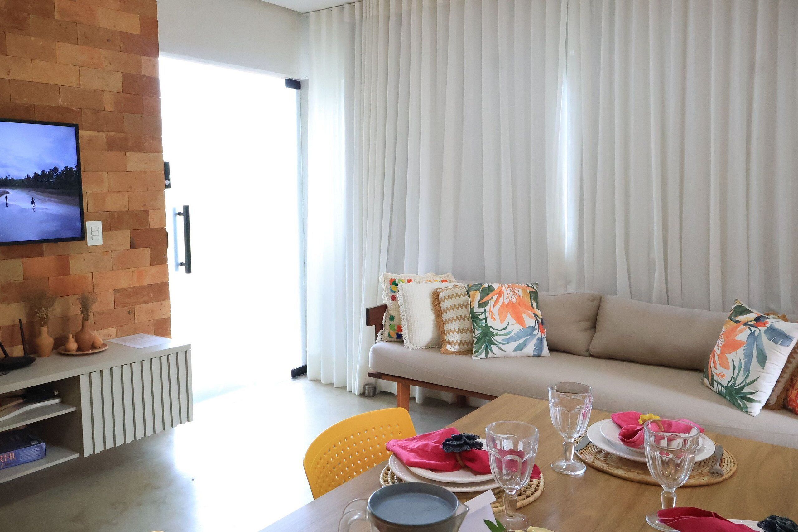 A living room with a sofa, a television mounted on a brick wall, a dining table with chairs, and a large window with curtains.