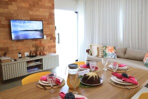 Un comedor con una televisión montada en una pared de ladrillo, un sofá con cojines de colores, y una mesa dispuesta con platos, vasos y un pastel.