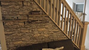Ein Wohnzimmer mit Steinwand, einer Holztreppe, einem dunkelgrauen Sofa mit Kissen und einem kleinen Regal mit Modellbauten.
