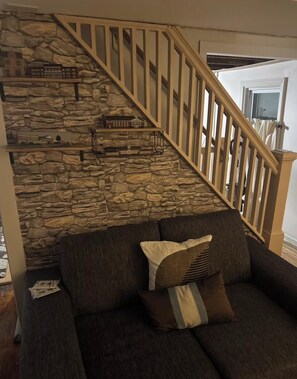 Un salon avec un mur en pierre, un escalier en bois, un canapé de couleur sombre agrémenté de coussins et une petite étagère où sont exposés des modèles de bâtiments.