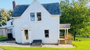 Ein weißes Haus mit dunklem Dach, einer Veranda und einem kreisförmigen Garten mit drei Schwein-Statuen.