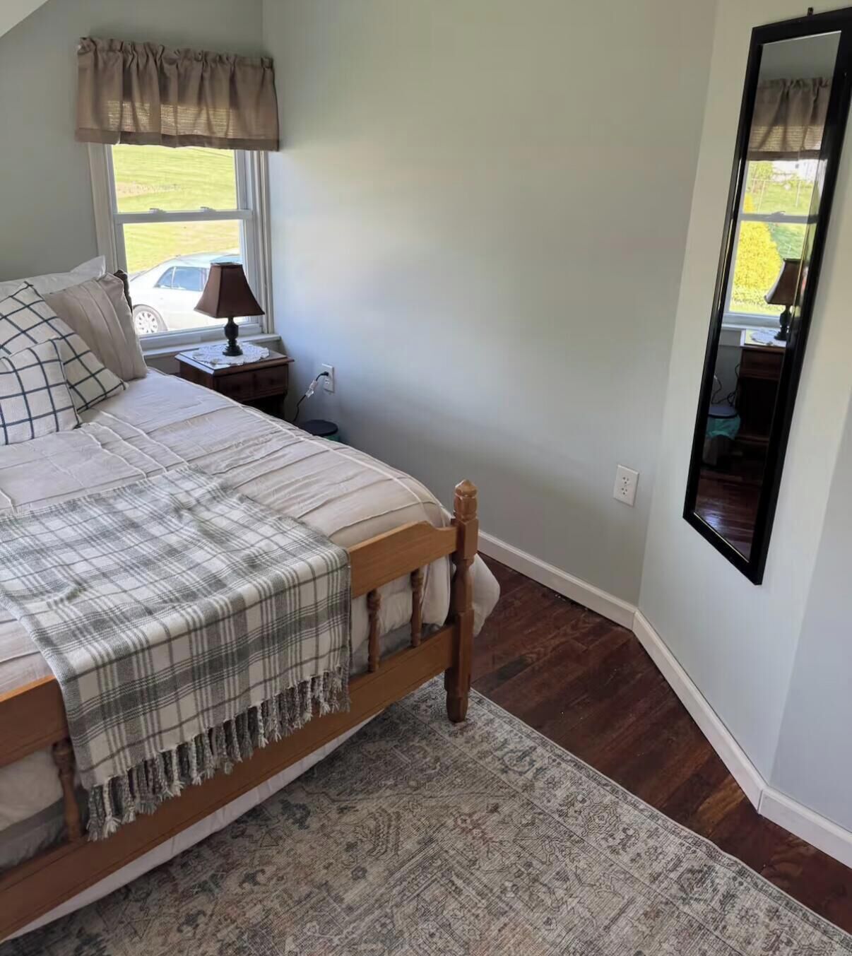 A bedroom with a bed, a nightstand, a lamp, a window with a view, and a mirror.