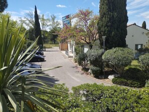 A parking lot with several cars, a building with a sign, and various trees and shrubs. - Campanile Arles (Arles)
