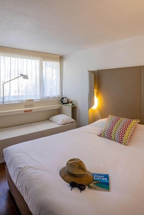 A hotel room with a bed, a bedside table with a lamp, a window with curtains, a headboard with a light, and a hat and book on the bed. - Campanile Arles (Arles)